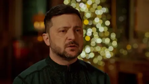 Reuters/Ukrainian Presidential Press Service Zelensky wearing a green shirt speaks in a dimly-lit room. An out-of-focus Christmas tree is in the background.